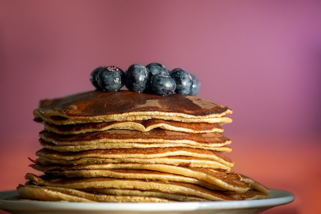 A tall stack of golden pancakes topped with fresh blueberries, drizzled with syrup, and served on a white plate, creating a mouthwatering breakfast scene