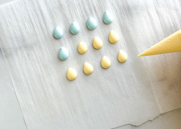 Image of Make your royal icing transfer eggs. These take 6-8 hours to dry, and can be made up to two weeks prior to assembling the cookies. Make sure your parchment paper is laying flat and select a location where the royal icing transfers will be able to lay and dry undisturbed.  Using the light blue and light yellow outline consistency icing, pipe small egg shapes onto the parchment paper. Use your scribe tool or toothpick to gently shape the eggs until desired shape is reached.  Allow the transfers to fully dry for 6-8 hours at room temperature, until the easily slide off the paper. You can save these in a sealed container for up to two weeks. 