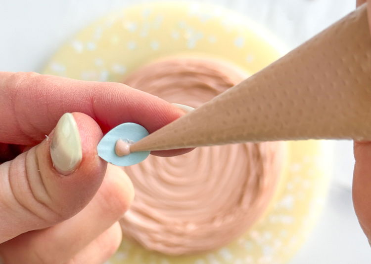 Image of Once the transfers are completely dry and easily slide off the parchment paper, carefully pick them up and add a small dot of light brown outline consistency icing onto the back of the transfer.  Carefully place each egg around the center of the cookie, pressing gently to secure them in place.  