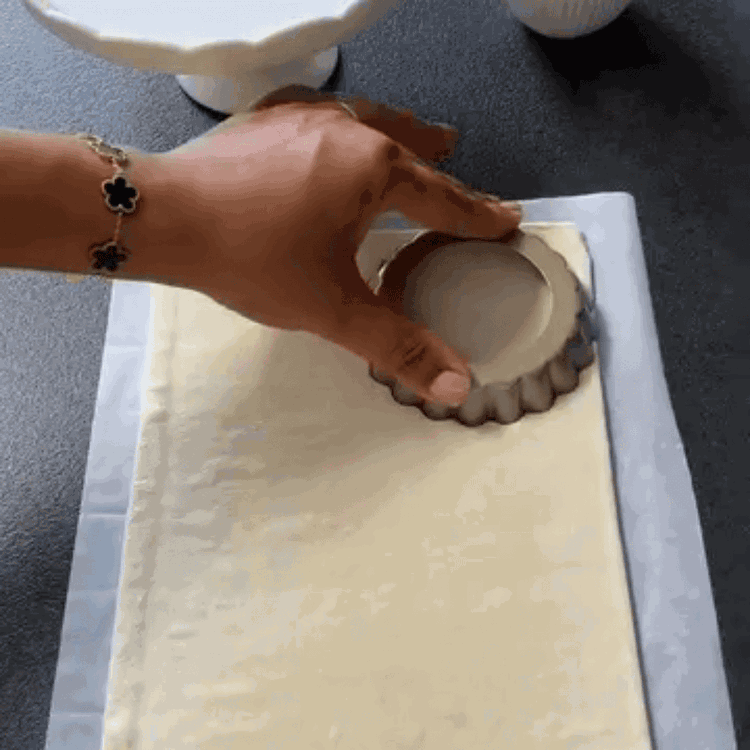 Image of Milföy hamurunu açın ve tartalet kalıplarını kesici olarak kullanarak 6 eşit daire kesin. Hamur dairelerini dikkatlice yağlanmış kalıplara yerleştirin, ortadan dışa doğru nazikçe bastırarak yükseltilmiş bir kenar oluşturun. Alt kısmı, pişirme sırasında kabarmasını önlemek için bir çatal ile birkaç kez...