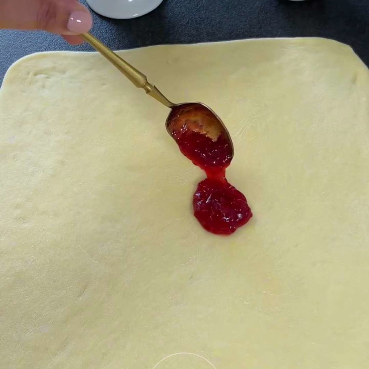 Image of Estira la masa en un rectángulo delgado y corta en aproximadamente 12 tiras iguales. Unta mermelada de fresa en cada tira y enrolla firmemente en espiral.