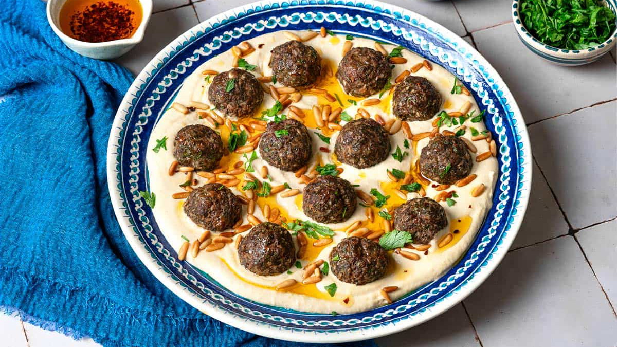 baked meatballs over hummus garnished with olive oil, pine nuts and parsley on a plate next to a blue napkin, a stack of blue plates with forks, and small bowls of parsley, pine nuts and olive oil.