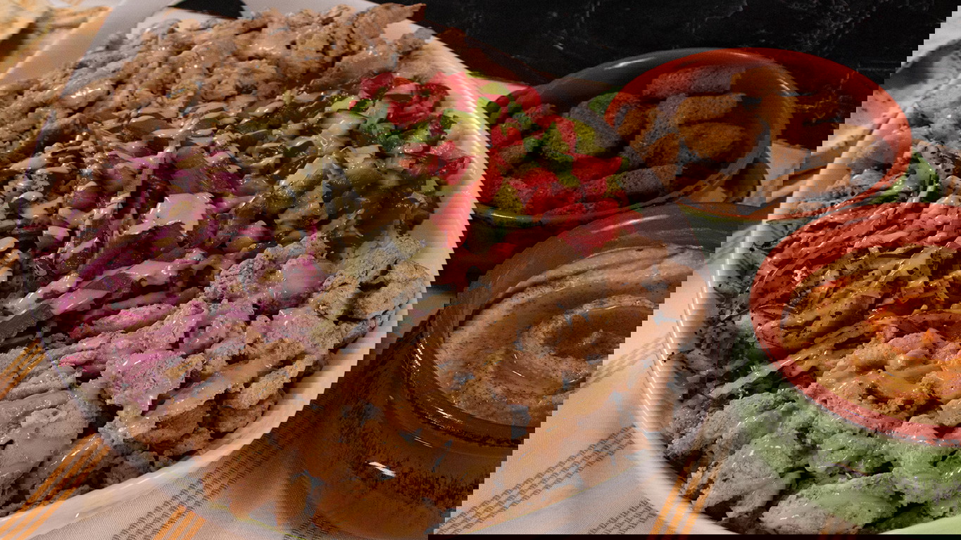 A large white dish filled with juicy pieces of Halal chicken, fresh cucumber and grape tomato salad, seasoned red onion, and Arabic pickles served over jasmine rice. Served next to two green dishes of falafel and hummus.