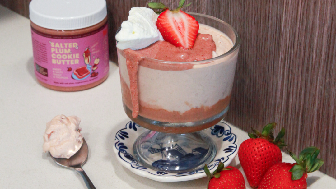 Clear glass with bottom layer of dark pink Salted Plum Cookie Butter. Rest of glass is filled with a fluffy light pink mousse, topped with more nut butter, a dollop of whipped cream, and a sliced strawberry. A jar of Rooted Fare's Salted Plum Cookie Butter is behind the glass and three bright red strawberries are on the table.