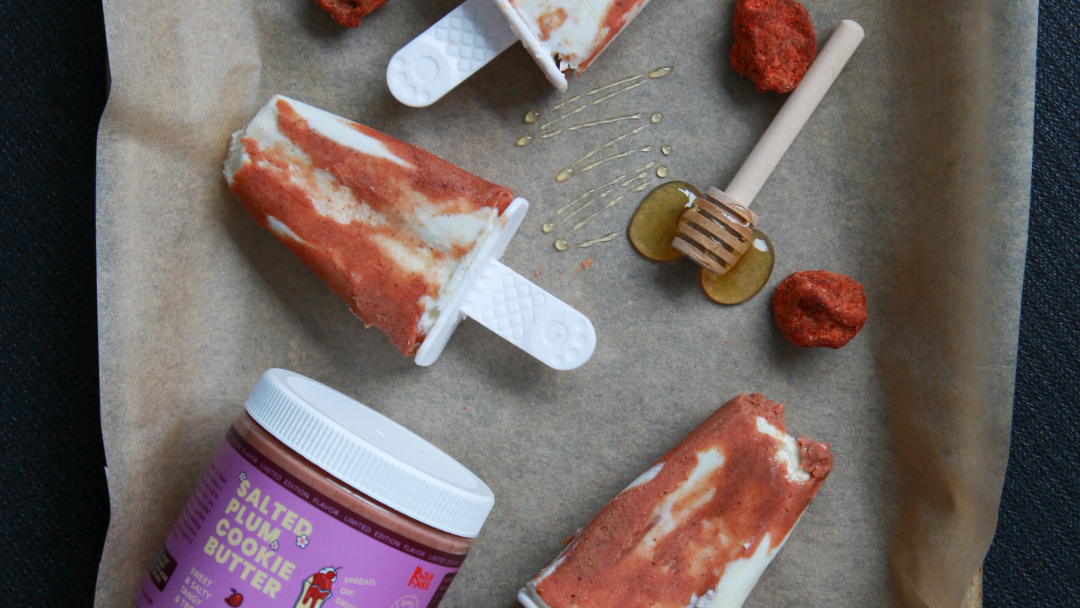 Sheet tray lined with parchment paper with three popsicles and ingredients on top. Popsicles have a creamy white base with burnt orange marbling made with Rooted Fare's Salted Plum Cookie Butter.