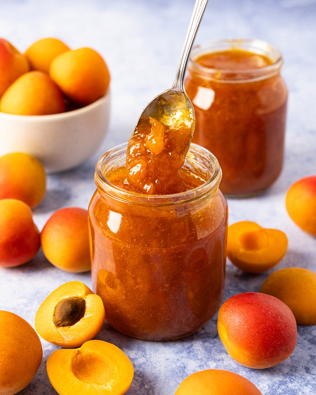 A jar of homemade apricot jam with a spoon being dipped in, surrounded by fresh apricots