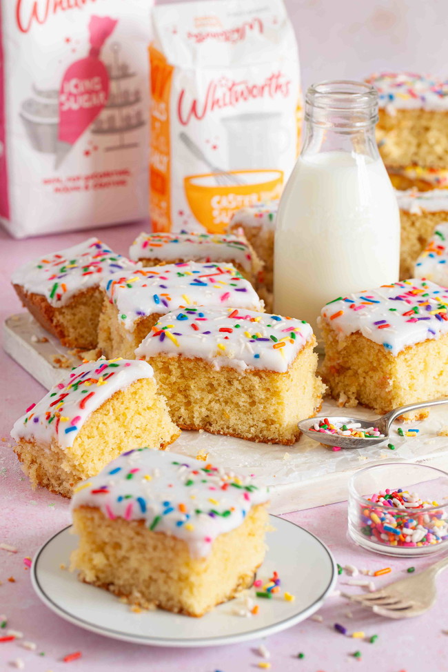 Slices of old school cake on a plate next to a bottle of milk and Whitworths caster and icing sugar