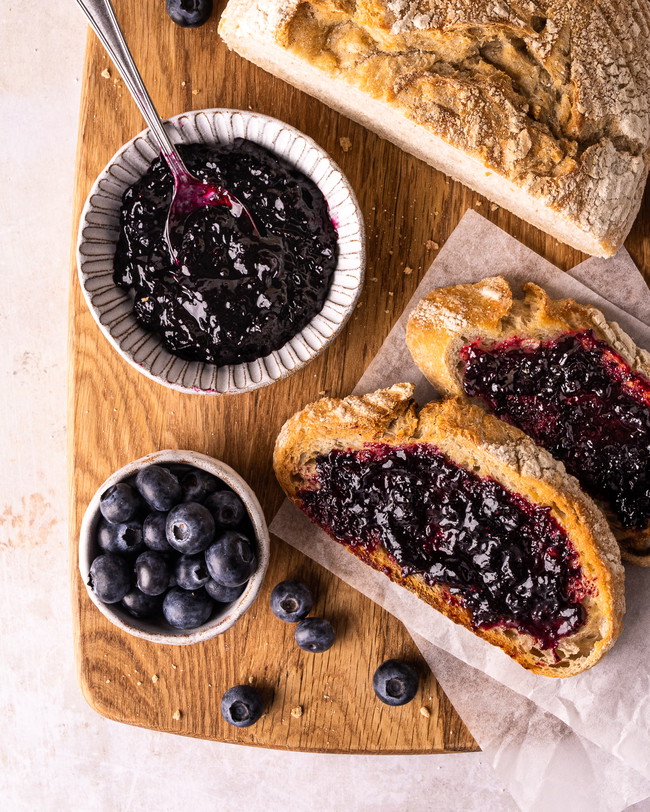 a board of toast with homemade blueberry jam, next to a jar of fresh jam and blueberries