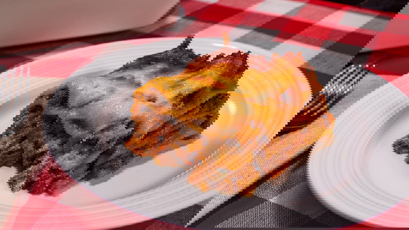 A slice of lasagna, with layers of Halal meat sauce, cheese filling, and tender lasagna noodles all topped with golden brown melted cheese. Served on a white plate on a red and white checkered tablecloth.