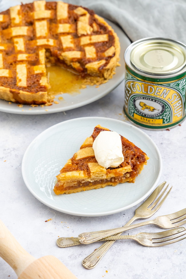 A slice of Lyle’s Easy Treacle Tart with a golden crust, topped with cream, next to a tin of Lyle's Golden Syrup 454g Tin. The full tart can be seen in the background oozing with delicious golden syrup.