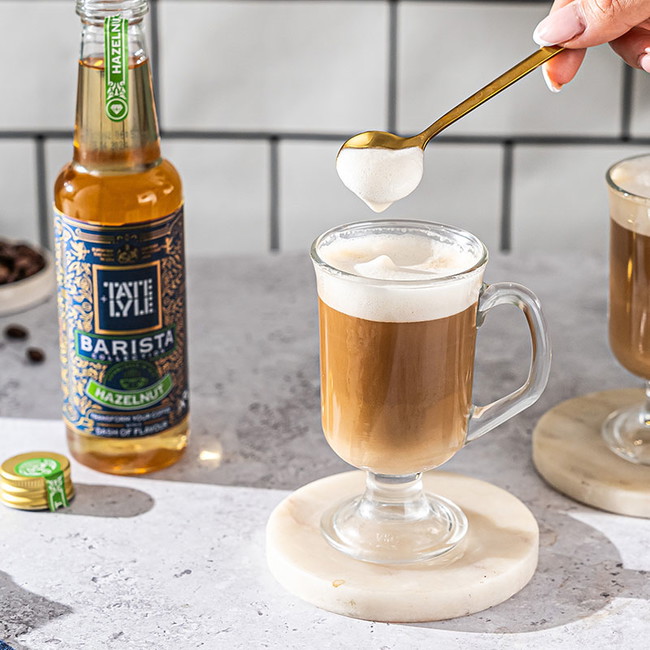 In a bright, artisanal kitchen, a hand carefully adds frothed milk into a hazelnut latte, accompanied by a bottle of Tate & Lyle Hazelnut Syrup.
