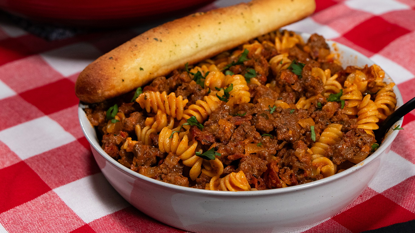 A white bowl with rotini pasta in a meaty red sauce with chunks of Halal ground beef, bacon, and pepperoni. Served with a breadstick on a red and white checkered tablecloth.