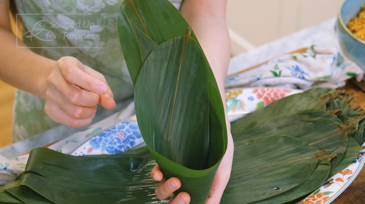 Image of Partially stack 2 bamboo leaves together with the smooth side facing up. Then, fold it slightly off the center into an ice cream cone. Make sure to over-lab the corner so the rice will not fall out.