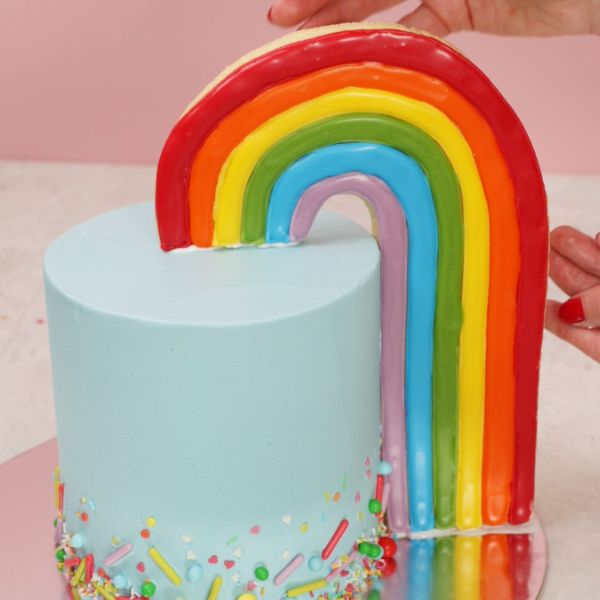 Image of Stelle den fertigen Regenbogenkeks vorsichtig in die Torte.💡 Fixiertipp: Verwende einen Klecks Royal Icing, um den Keks an der Torte sowie an der Tortenplatte zu befestigen – das gibt zusätzlichen Halt.
