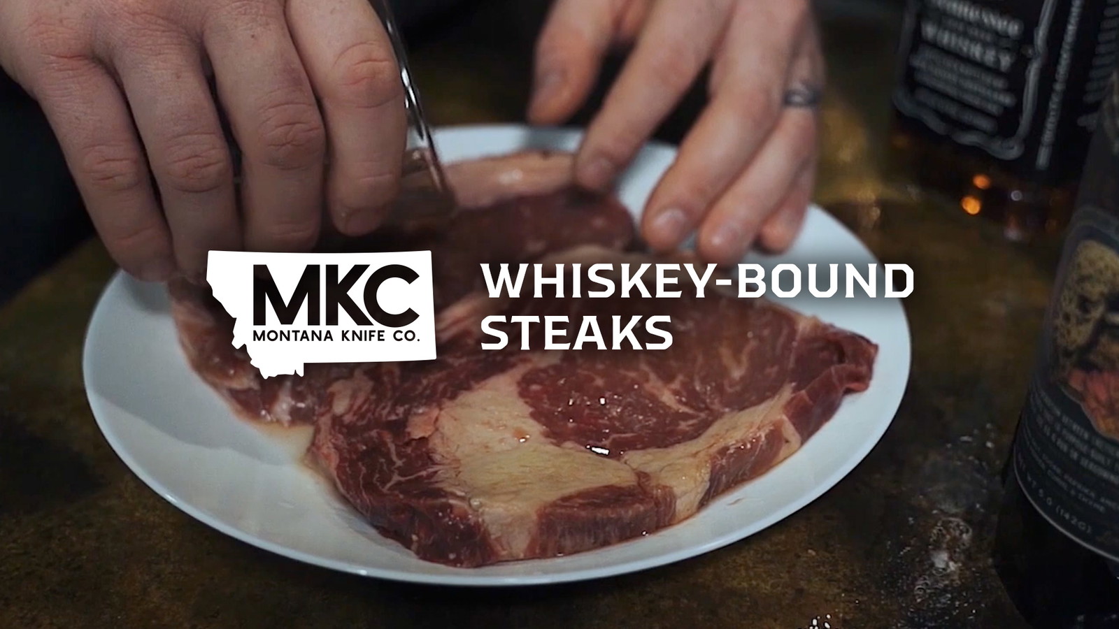 Hands using a knife to prepare raw ribeye steaks on a white plate with whiskey bottle in the background.