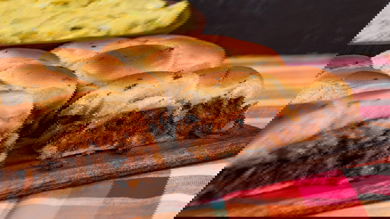 Sliders made with tender and juicy BBQ pulled beef, topped with crispy onions and melted cheese and served on a black serving board. Behind it is a wooden bowl of creamy homemade potato salad. 