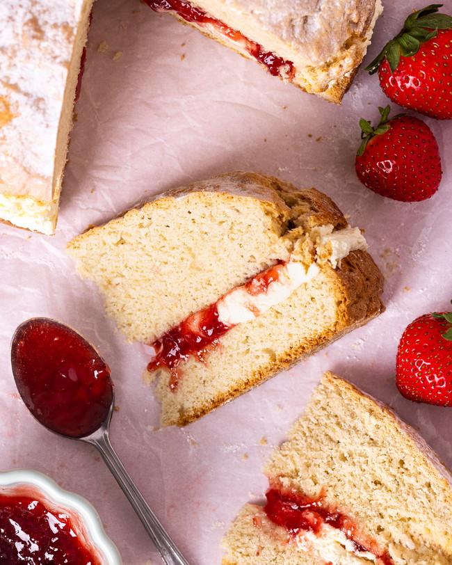 A slice of giant scone cake filled with clotted cream and strawberry jam, surrounded by fresh strawberries and a spoonful of jam on parchment paper.