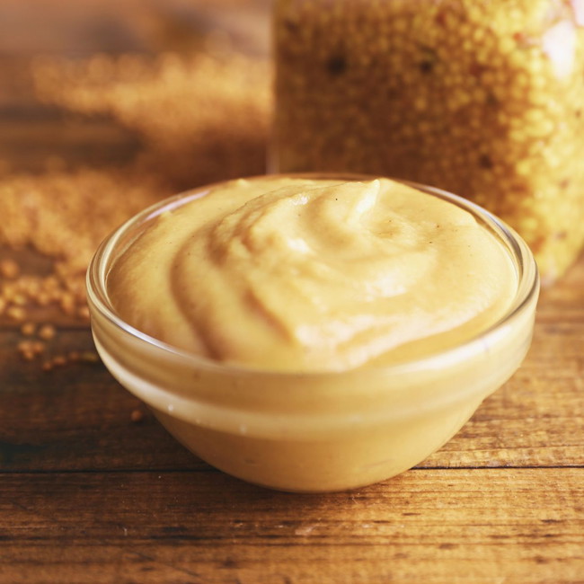 Small glass bowl filled with creamy spicy maple mustard dipping sauce, set on a rustic wooden surface with mustard seeds and a jar of whole grain mustard in the background.