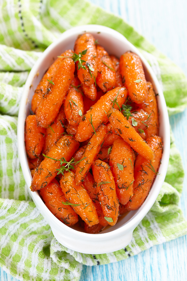 White dish filled with roasted baby carrots glazed with maple syrup and garnished with fresh herbs, placed on a green-striped towel over a blue wooden table.