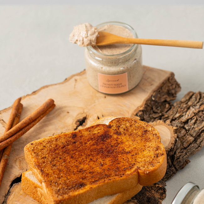Jar of cinnamon maple whipped butter made with Wild West Waffles Cinnamon Maple Syrup, with a wooden spoon resting on top, placed on a rustic wood slab next to toasted cinnamon sugar bread and whole cinnamon sticks.