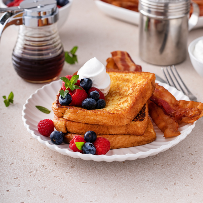 Stack of vanilla maple French toast topped with whipped cream and fresh berries, served with crispy bacon on a white plate and drizzled with Wild West Waffles Vanilla Maple Syrup.