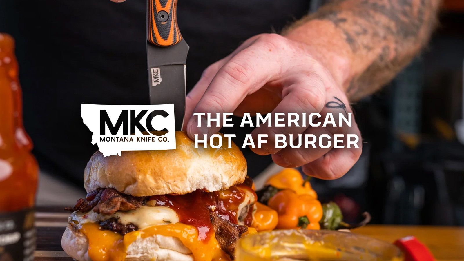 A close-up view of a man’s tattooed hands holding a burger with a knife stuck into it.