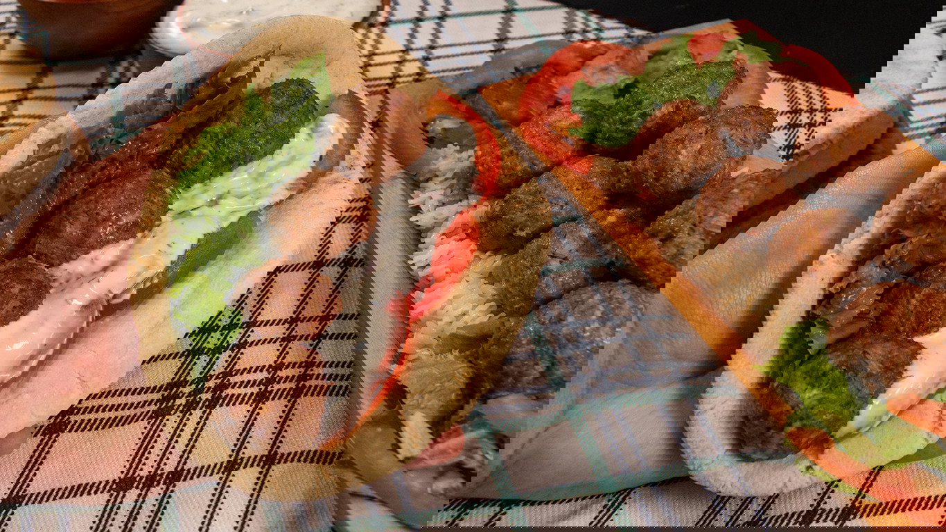 Golden-brown lamb meatballs, served on a pita with tomatoes, lettuce, and tzatziki sauce alongside a wooden platter with rice, veggies, and topped with more meatballs.