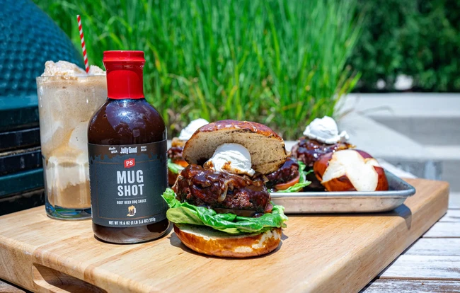 Root Beer Float Burger made with Mug Shot Root Beer BBQ Sauce on a pretzel bun, topped with caramelized onions and scooped goat cheese, served alongside a root beer float and a bottle of PS Mug Shot BBQ Sauce on a wooden board outdoors.