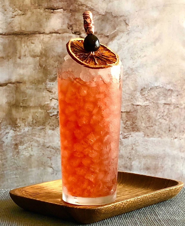 Red huged cocktail in a tall glass filled with crushed ice and a dried orange slice garnish.
