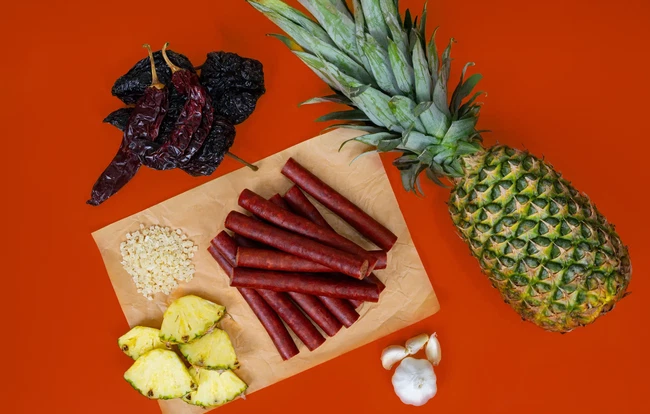 Al Pastor Snack Sticks made with PS Seasoning No. 854 Al Pastor Snack Stick Seasoning, surrounded by dried guajillo and ancho chiles, fresh pineapple, garlic cloves, and diced pineapple on an orange background.