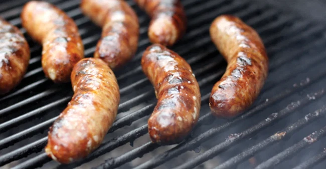 A close-up view of several sausages being grilled on a barbecue. The sausages are evenly spaced across the metal grates, with a browned and slightly charred exterior that suggests they are being cooked over high heat. Wisps of smoke rise from the grill, adding to the sense of sizzling heat and outdoor cooking