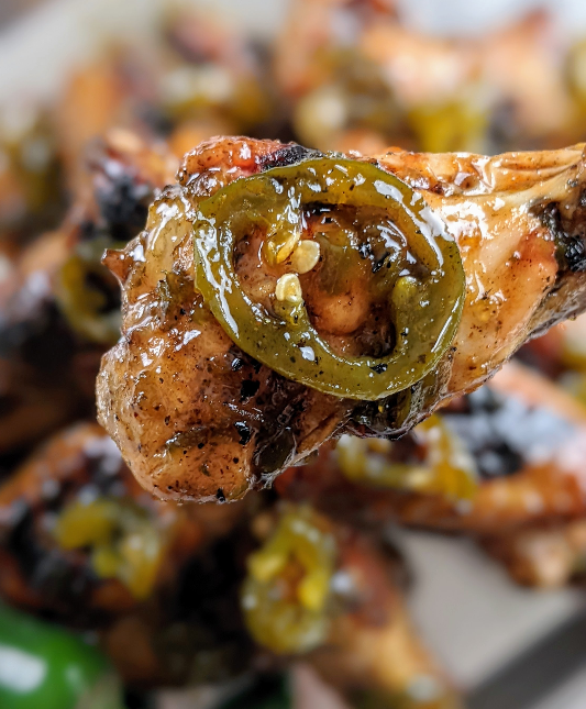 A close-up, vertical shot of a single Candied Jalapeno Wing from PS Seasoning, held up to the camera. The wing is dripping with a glossy, sweet and spicy glaze, and a large slice of bright green candied jalapeño sits perfectly on top, showcasing the texture and detail of the recipe.