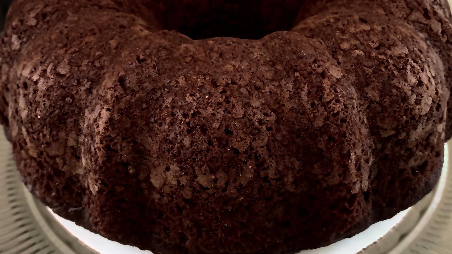 A whole, dense chocolate pound cake with a deep cocoa color, displayed on a simple cake stand for an elegant presentation.