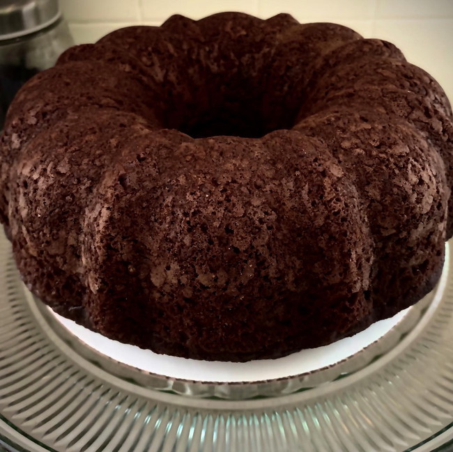 A whole, dense chocolate pound cake with a deep cocoa color, displayed on a simple cake stand for an elegant presentation.