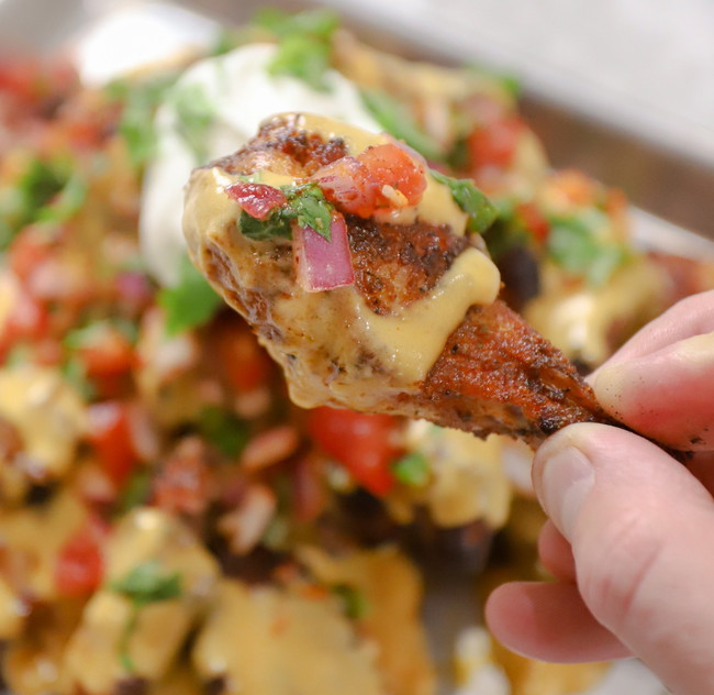 A hand holds up a single chicken wing covered in melted cheese sauce, diced red onion, tomatoes, and cilantro. The rest of the tray of Nacho Wings is visible in the background, out of focus. These nachos are seasoned with PS Seasonings El Jefe Adobo rub. 