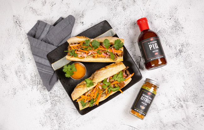Two toasted baguette banh mi sandwiches filled with shaved beef seasoned in PS Seasoning Sesame Chili Citrus Wing Rub, quick-pickled vegetables, cilantro, and creamy Notorious PIG BBQ mayo, served on a black plate with dipping sauce. shaved beef banh mi recipe, BBQ banh mi sandwich, PS Seasoning Sesame Chili Citrus, Notorious PIG BBQ Sauce sandwich, pickled vegetable banh mi, sweet and spicy BBQ banh mi, Vietnamese BBQ sandwich recipe