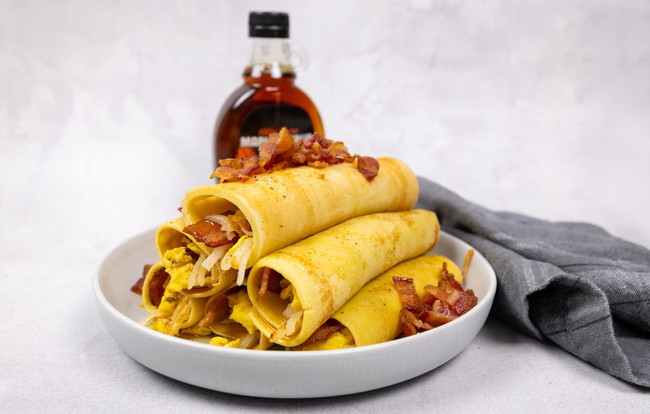 Three rolled breakfast crêpes filled with scrambled eggs, crispy bacon, and hash browns, seasoned with PS Seasoning Chicken and Waffle Wing Rub, served on a white plate with maple syrup in the background. bacon breakfast crepe recipe, chicken and waffle seasoning breakfast, PS Seasoning crepe rolls, savory breakfast crepe with eggs and bacon, maple bacon breakfast wrap, brunch crepe recipe, breakfast meal prep ideas