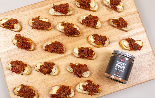 Toasted crostini topped with creamy brie cheese and smoky bacon jam made with PS Seasoning Cherry Bomb Sweet Heat BBQ Rub, arranged on a wooden board. bacon jam brie crostini, sweet heat BBQ appetizer, PS Cherry Bomb seasoning recipe, bacon jam with brie, easy holiday appetizers, crostini with brie and bacon, sweet and savory party bites