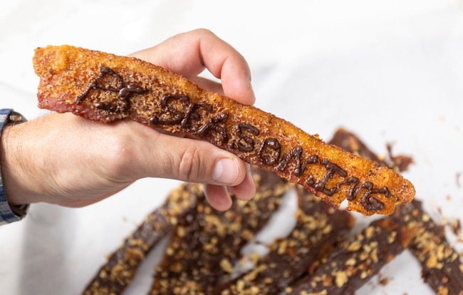 Hand holding a strip of chocolate-covered bacon seasoned with PS Seasoning Churro Dust BBQ Rub, topped with piped chocolate lettering and chopped nuts, with more bacon strips in the background. chocolate covered bacon recipe, churro dust bacon, PS Seasoning sweet and savory snack, BBQ spiced dessert, cinnamon sugar bacon treat, sweet and salty bacon, candied bacon with chocolate