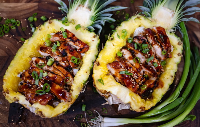 Grilled teriyaki chicken thighs seasoned with PS Seasoning Big Kahuna BBQ Rub, drizzled with Big Kahuna Pineapple Teriyaki BBQ Sauce, served over jasmine rice inside carved pineapple halves and topped with sesame seeds and green onions. teriyaki chicken pineapple bowls, Big Kahuna BBQ Rub recipe, pineapple teriyaki chicken, PS Seasoning island-inspired dinner, grilled chicken pineapple boat, summer BBQ chicken recipe, Hawaiian style chicken and rice