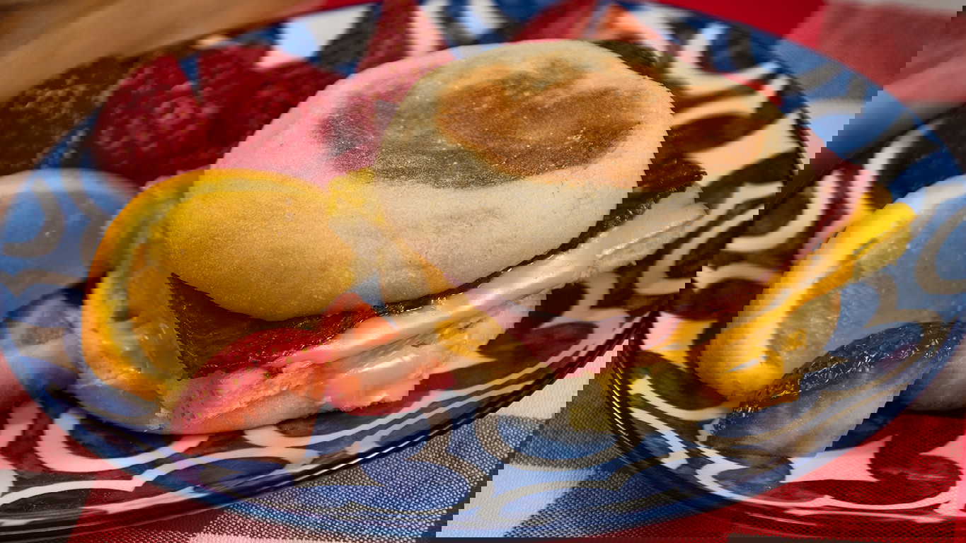 A breakfast sandwich with a slice of scrambled egg, hashbrown, and spinach, topped with melty cheese and Halal beef strips on a toasted English muffin. Served with fruit on a blue patterened plate.