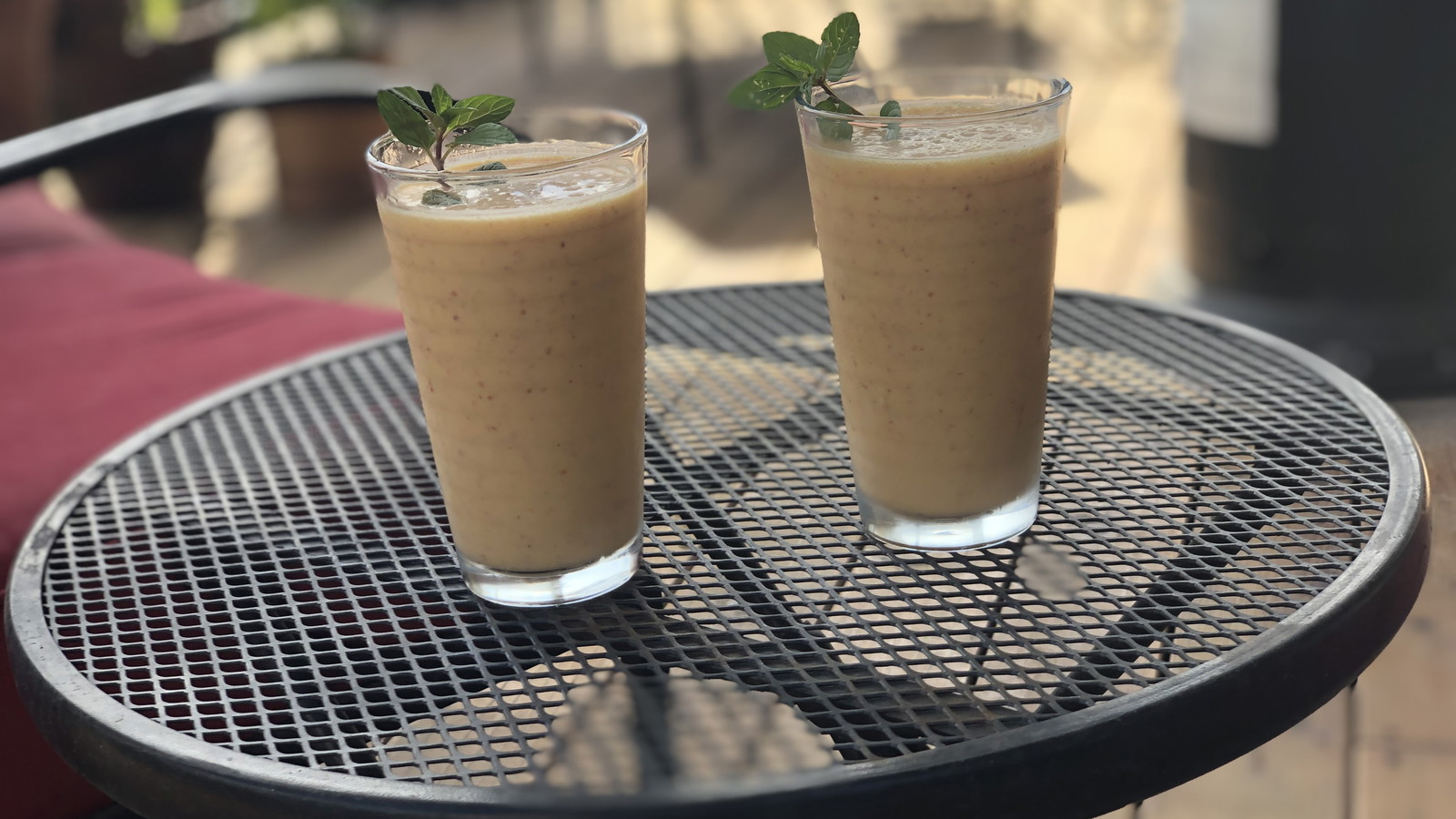 Two glasses of peach lassi garnished with fresh mint, served on a black patio table in natural sunlight