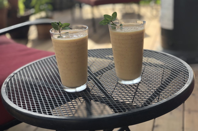 Two glasses of peach lassi garnished with fresh mint, served on a black patio table in natural sunlight