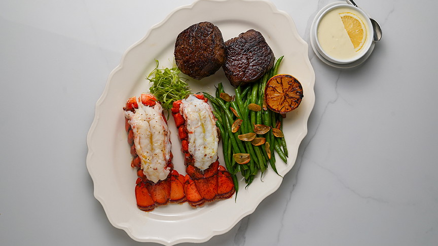 Two Lobster tails and two steaks on a white plate with veggies