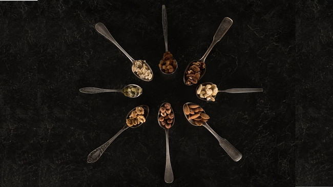 Assorted raw nuts displayed in vintage spoons arranged in a circle on a dark slate background including macadamias, pecans, almonds, cashews, walnuts, and hazelnuts