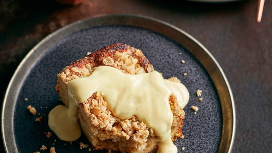 Close-up of a golden, crumbly apple crumble with cinnamon-spiced apples, topped with creamy custard being poured over it.