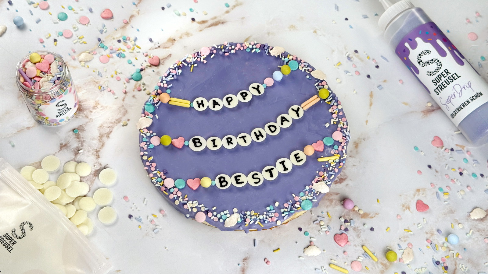 Cheesecake mit lavendelfarbenem SuperDrip überzogen, bestreut mit StreuselMix PastellKarussell und dekoriert mit weißen SuperDrops, die mit schwarzen Buchstaben wie Freundschaftsarmbänder angeordnet sind. Bunte Geburtstagstorte im Taylor-Style