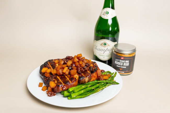 Grilled and smoked thick-cut pork chops topped with champagne cherry apple chutney, served over roasted asparagus. PS Seasoning Rodeo Rub and a bottle of champagne placed behind the plate. Sweet, smoky, and savory BBQ dinner recipe. smoked pork chops recipe, Rodeo Rub pork chops, cherry apple chutney, PS Seasoning Rodeo Rub, champagne chutney pork, smoked pork with fruit topping, BBQ pork chops with asparagus