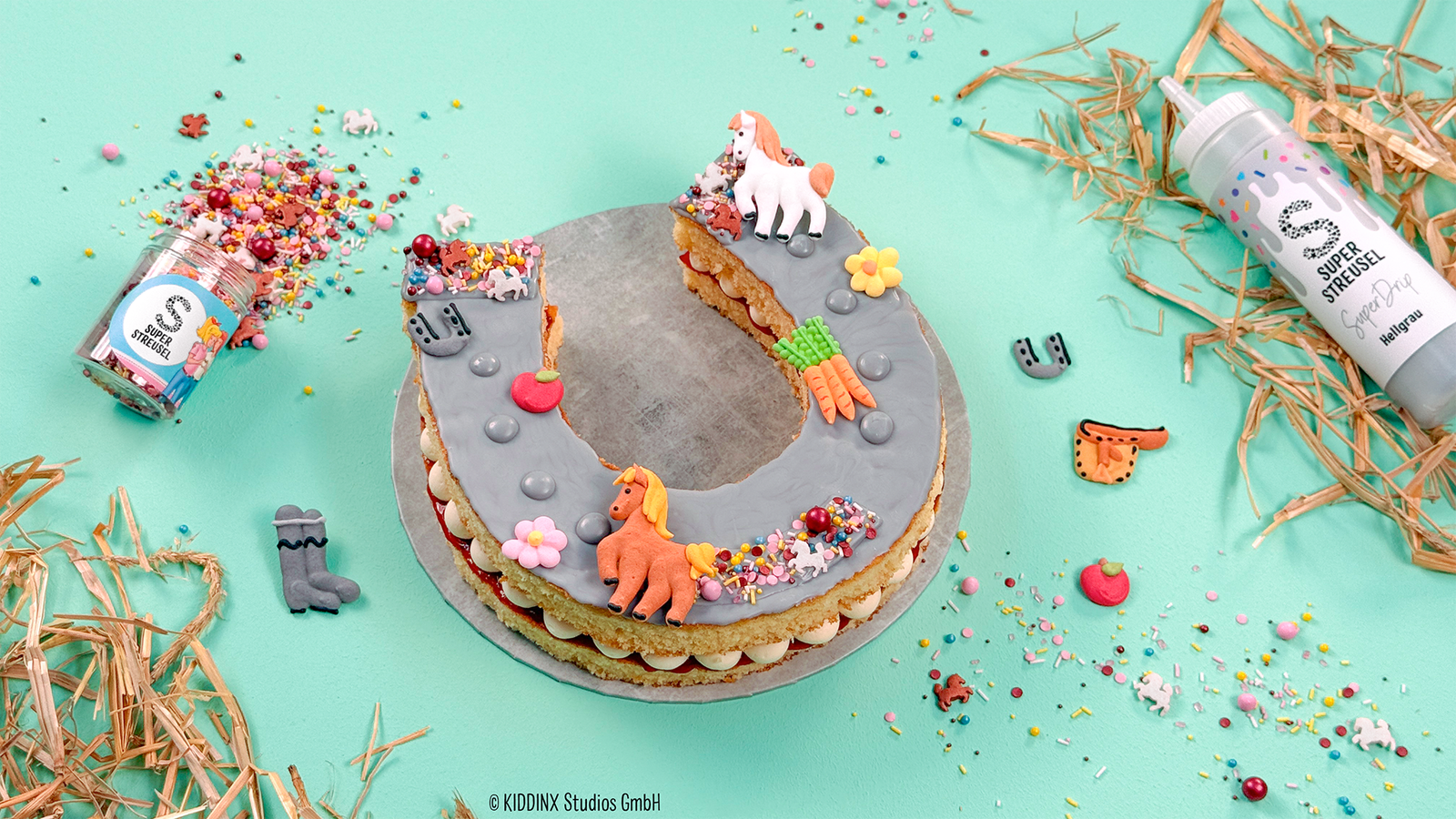 Bibi & Tina Hufeisen Letter Cake aus Vanille-Blechkuchen mit Vanille-Mascarpone-Frosting und Erdbeermarmelade, dekoriert mit hellgrauem SuperDrip, StreuselMix BibiUndTina und SuperDinger HoppeHoppeReiter. Pferde-Geburtstagskuchen in Hufeisenform.