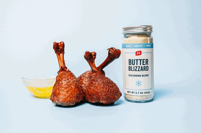 Three grilled chicken lollipops seasoned with PS Seasoning Butter Blizzard blend, arranged beside a bowl of melted butter and a jar of Butter Blizzard seasoning on a light blue background. Juicy, crispy chicken perfect for BBQ or game day.  SEO Keywords: Butter Blizzard chicken lollipops, PS Seasoning Butter Blizzard, grilled chicken drumstick recipe, BBQ chicken lollipop, garlic butter chicken rub, chicken lollipops on smoker, game day chicken recipe